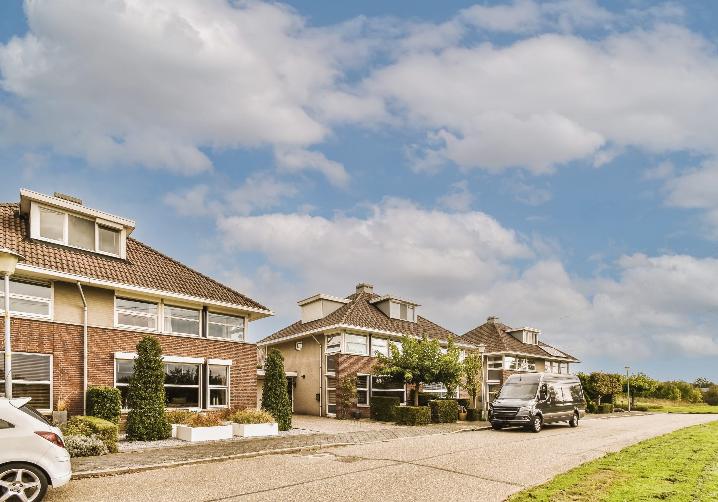 a house with a car parked in front of it some houses with cars parked on the side of the road and clouds in the sky above them are white fluffy clouds