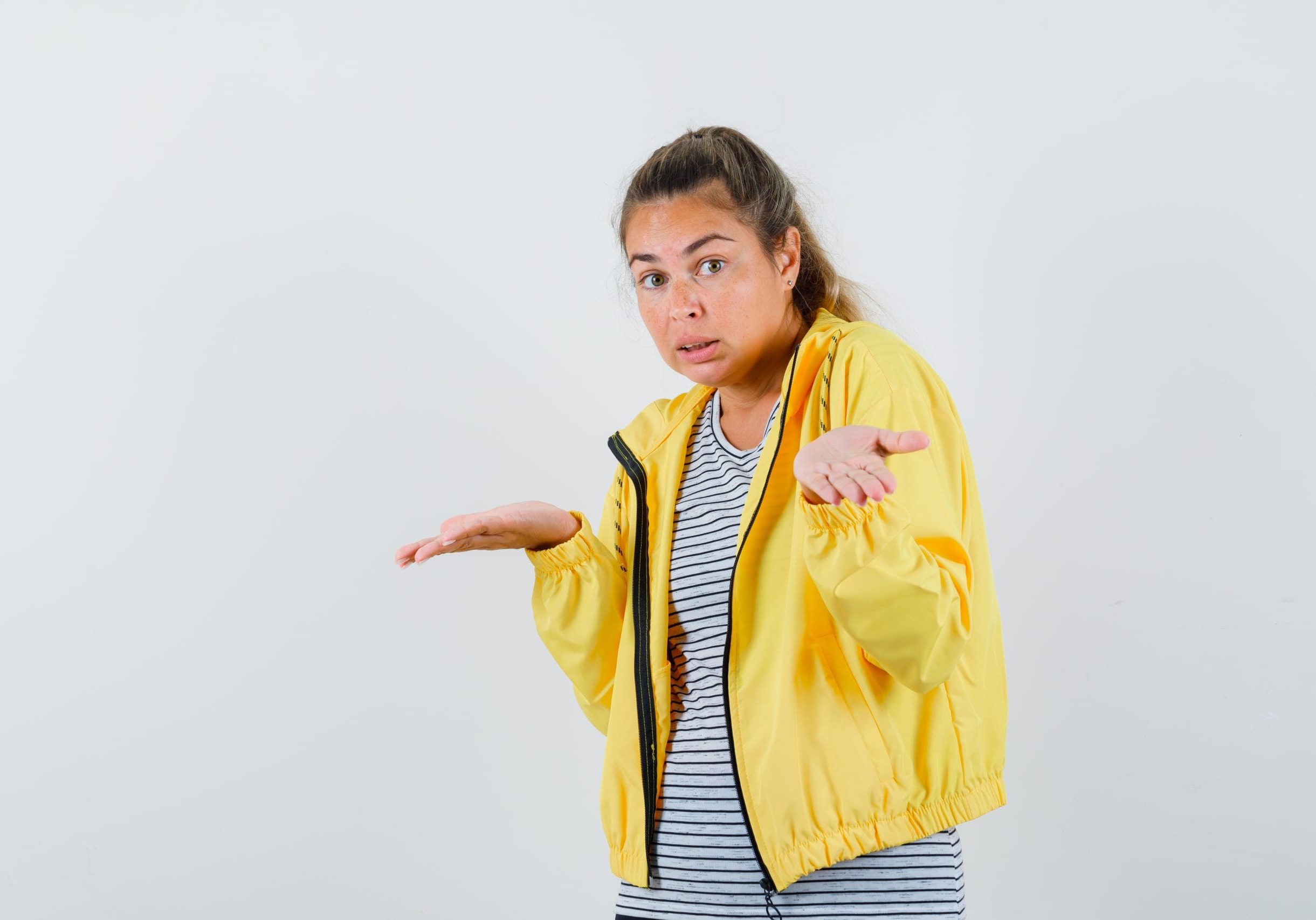 blonde woman stretching hands in questioning manner in yellow bomber jacket and striped shirt and looking pensive , front view. blonde woman stretching hands in questioning manner in yellow bomber jacket and striped shirt and looking pensive , front view.