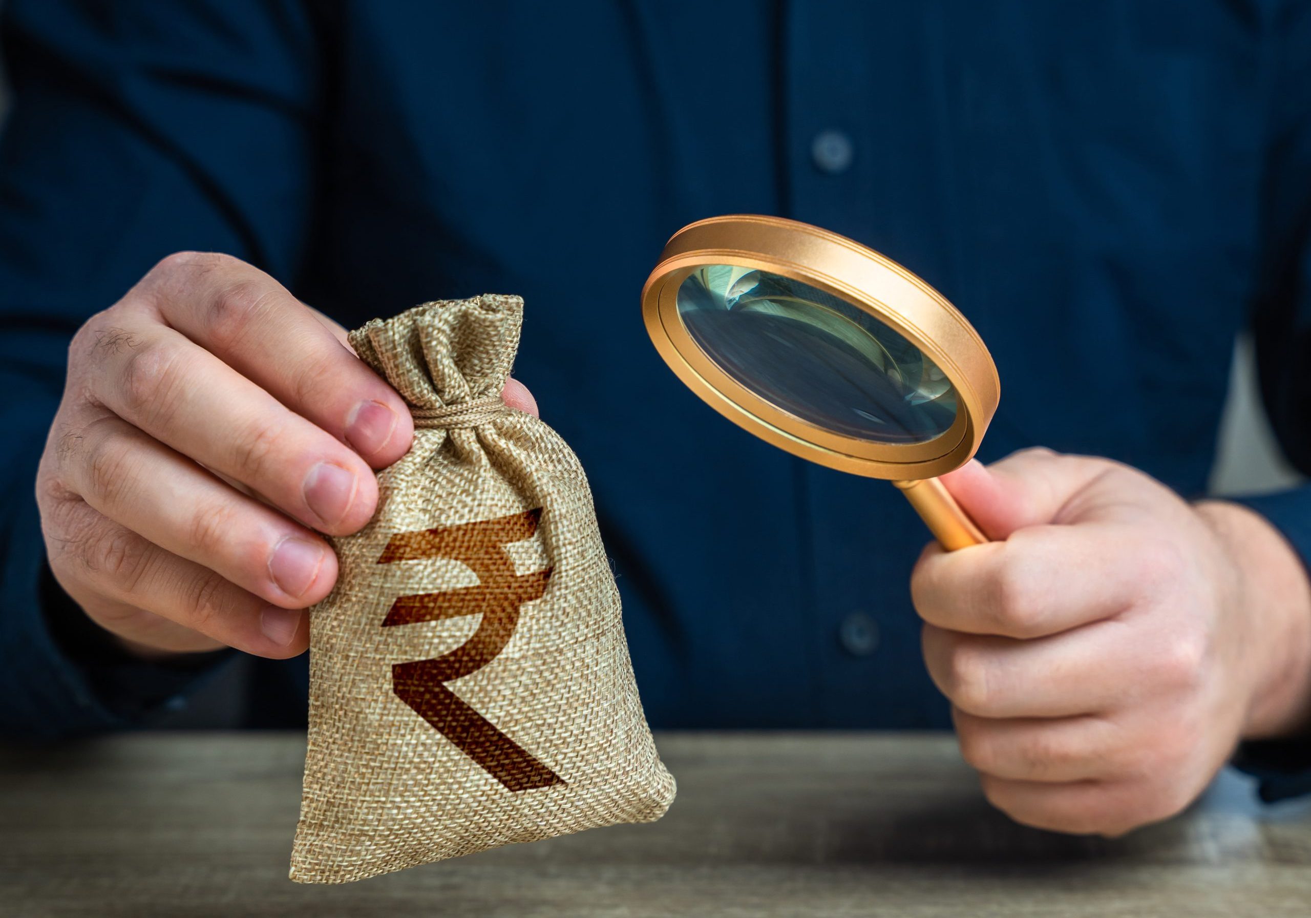 A man examines a indian rupee money bag through a magnifying glass. Investigating capital origins. Anti money laundering, tax evasion. Study terms and conditions on deposit or loan.